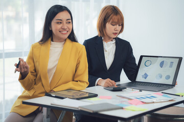 Confident Collaboration: Two businesswomen, clad in professional attire, engage in a discussion while reviewing financial data on a laptop.  One leans in with a thoughtful expression.