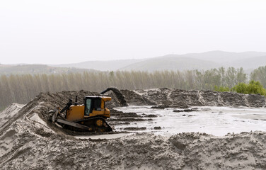 Industrial scene at a sand production plant. An excavator with a bucket is leveling a large pile of...