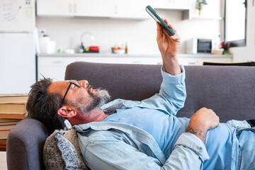 Obraz premium Mature adult man comfortably reclining on the couch at home, wearing glasses and a beard, using a smartphone to scroll through social media and checking notifications and emails for leisure 