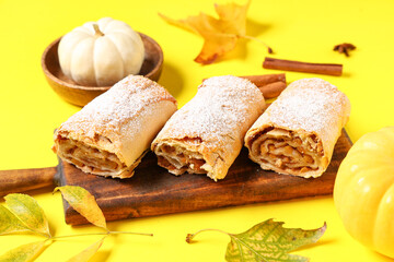 Wooden board with cut tasty pumpkin strudel, cinnamon and autumn leaves on yellow background