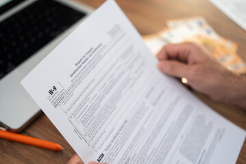 Obraz premium Consultant looks over W-9 tax form to provide TIN to client for reporting consulting fees already paid during year at wooden table with euro bills and laptop during break