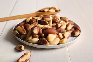 Plate and spoon with tasty Brazil nuts on white wooden background