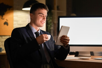 Young businessman with coffee and tablet computer working in office at night