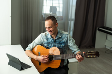 Portrait of man taking online guitar lesson looking at laptop screen. Retired male learning to play guitar watching webinar on computer at home
