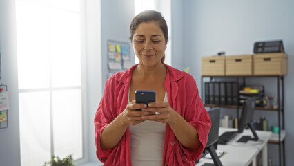 Woman smiling while using smartphone in modern office with organized workspace and bookshelf in background