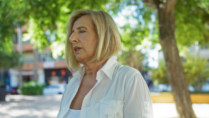 Beautiful mature woman with blonde hair and short hairstyle standing outdoors in an urban park setting with greenery and city buildings in the background