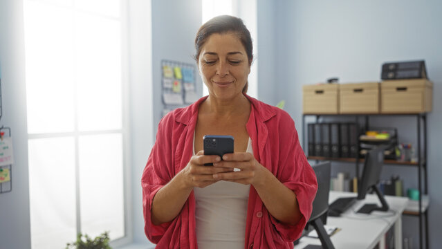 Woman smiling while using smartphone in modern office with organized workspace and bookshelf in background