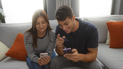 Man and woman using smartphones and credit card at home, sitting on a comfortable couch, enjoying their time together in a modern, cozy living room interior.