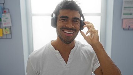 Handsome man smiling indoors in casual attire wearing headphones in an office setting with shelves and papers in the background