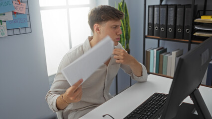 Young man in office fanning himself with documents while sitting at desk surrounded by binders and...