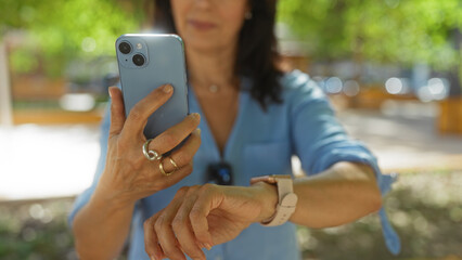 Middle-aged woman using smartphone in an urban park while checking her smartwatch on a sunny day outdoors