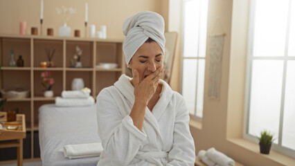 Woman yawning in a spa room wearing a white robe with a towel on her head, surrounded by a serene interior with organized shelves and natural light coming through the windows.