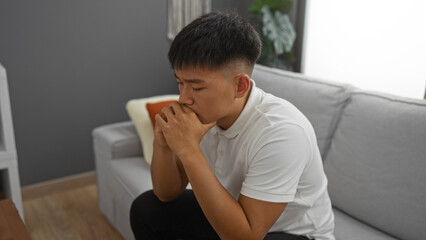 Young man sitting indoors in a modern living room, appearing pensive and reflective, wearing a white shirt and surrounded by minimalist decor