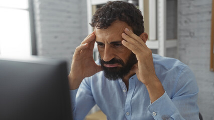 Young hispanic man with a beard sitting in an office with a stressed expression, hands on temples, focused on a computer screen in an indoor professional workplace