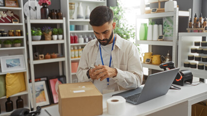 Young man with beard using mobile in home decor store, surrounded by shelves filled with various decorative items, scanning product at counter, laptop and barcode scanner visible.