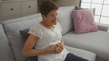 Elderly hispanic woman with stomach pain sitting on a couch in a living room, wearing glasses and a white blouse, showcasing a home interior setting.