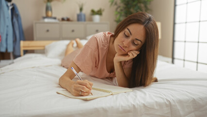 Young woman lying on a bed in a bedroom, writing in a notebook with a focused expression, surrounded by indoor plants and home decor.