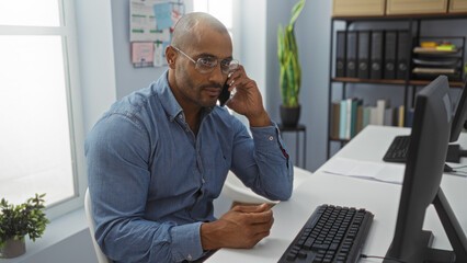 Fototapeta premium Handsome african american man working in an office while talking on the phone, surrounded by various documents and shelves filled with files, showcasing a focused and professional demeanor