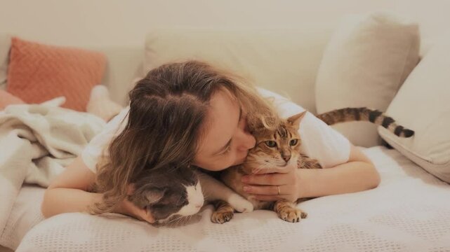 happy woman lying on sofa hugging two cute cats, joyfully kissing bengal cat and non-pedigree fluffy cat. Joyful family person friends with animals pets concept love care. girl casual clothes pajamas