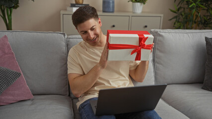 A handsome young hispanic man in a living room apartment, holding a gift box with a red ribbon, smiling while sitting on a gray couch with a laptop on his lap.