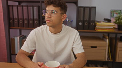 Young hispanic man with glasses sitting in an office holding a cup, surrounded by shelves with...