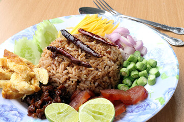 Rice mixed with shrimp paste consisting of sweet pork, chinese sausage, fried eggs, cucumber slice, sour mango, shallot, yard long bean, roasted peppers and lemon. Placed with a spoon and fork.
