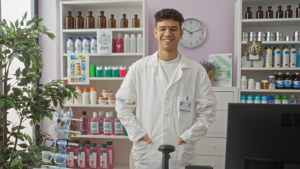 Fototapeta premium Hispanic male pharmacist in a white coat smiles confidently while standing in a well-organized pharmacy filled with various medical products and herbs.