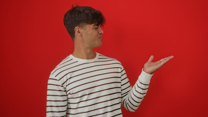 A young hispanic man with a striped shirt presents something with his extended hand against a red...