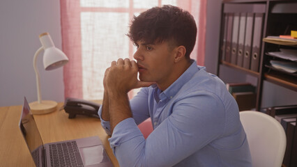 Handsome young hispanic man in an indoor office setting, deep in thought at a wooden desk with a...