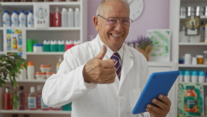 An elderly caucasian man with grey hair smiling and giving a thumbs-up while holding a tablet in a well-stocked pharmacy interior.