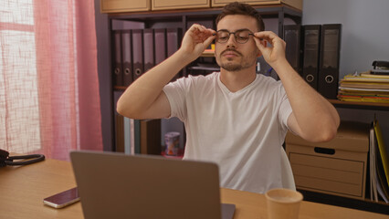 A handsome hispanic man with a beard, adjusting his glasses while sitting in an office with...