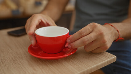 Young hispanic man enjoying coffee at a wooden table in a cozy indoor bakery, holding a red cup with both hands.