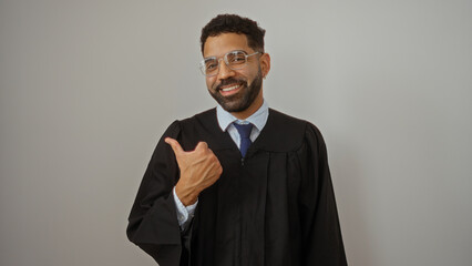 Young man in graduation gown smiling and pointing thumb over isolated white wall background