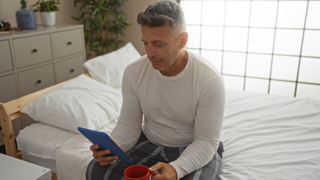 Middle-aged man sitting on bed in bedroom holding tablet and red mug while wearing casual clothes - Powered by Adobe