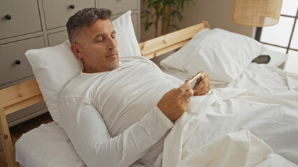 Middle-aged man with gray hair lying in a cozy bedroom, using a smartphone on a comfy bed under white sheets.