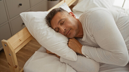 Middle-aged man sleeping peacefully in his bedroom, resting on a white pillow and dressed in light...