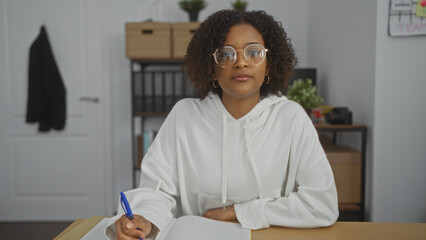 Young african american woman with curly hair in a white hoodie working in an office, sitting at a...