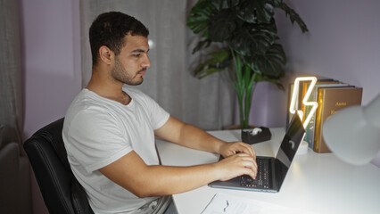 Young hispanic man working on laptop in modern living room with plants and neon lamp