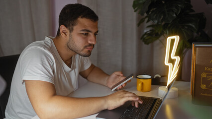 Hispanic man working on laptop at home with smartphone in hand, surrounded by cozy living room environment including books and a decorative lamp.