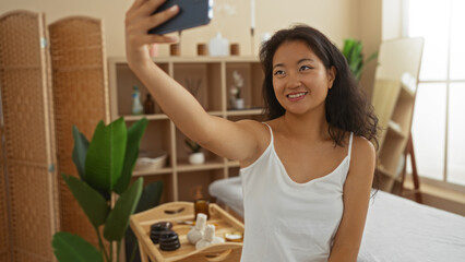 Woman smiling in a spa room taking a selfie, surrounded by natural decor and wellness items, capturing a relaxing indoor moment in a beauty center in china.