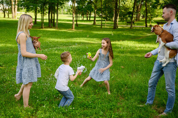 Fototapeta premium Children playing with soap bubbles in park. Boy and girl blowing soap bubbles from toys, happy parents standing nearby holding pets, dogs. Concept of happy childhood, summer holidays, family weekend