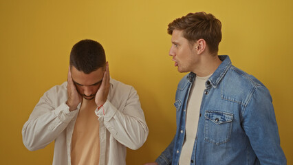 Two men argue as one covers his ears against a yellow wall, highlighting tension among adult male...