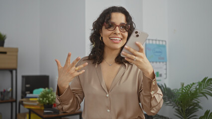 A young, brunette hispanic woman in an office enthusiastically talks on her smartphone, wearing...