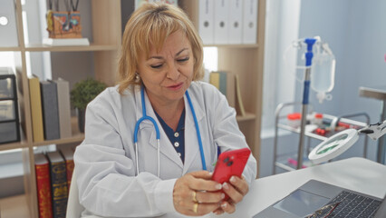 Mature caucasian female doctor using smartphone while sitting in a clinic office with medical equipment in the background.