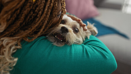 A young african american woman with braids holds and pets her happy dog in a cozy indoor living room setting.