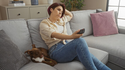 Beautiful young caucasian woman relaxing on a couch with her dog in a cozy living room, holding a remote control and looking thoughtful in a modern apartment.