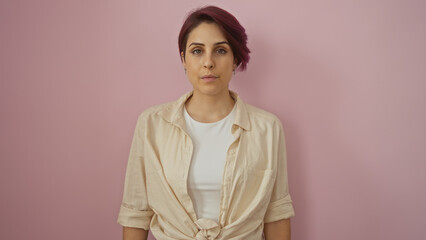 Portrait of a beautiful young woman with short hair standing against an isolated pink background wall, wearing a casual beige shirt and white top, looking confident and serene.