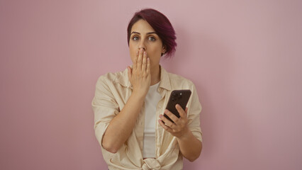 A young, beautiful, caucasian woman with short hair holds a smartphone while standing isolated against a pink wall, appearing surprised and covering her mouth with her hand.