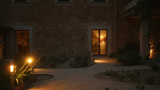 Fototapeta Exterior view of a cozy, illuminated stone house at night, showing glowing garden path lights, warm indoor lighting, and inviting decor, creating a serene and charming atmosphere.
