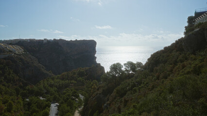 A scenic coastal landscape featuring cliffs overlooking the mediterranean sea, lush greenery, and distant houses under a clear sky.